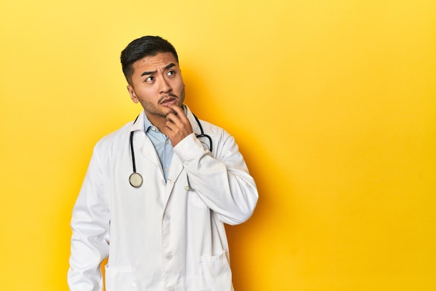 Asian doctor in white coat with stethoscope relaxed thinking about something looking at a copy space