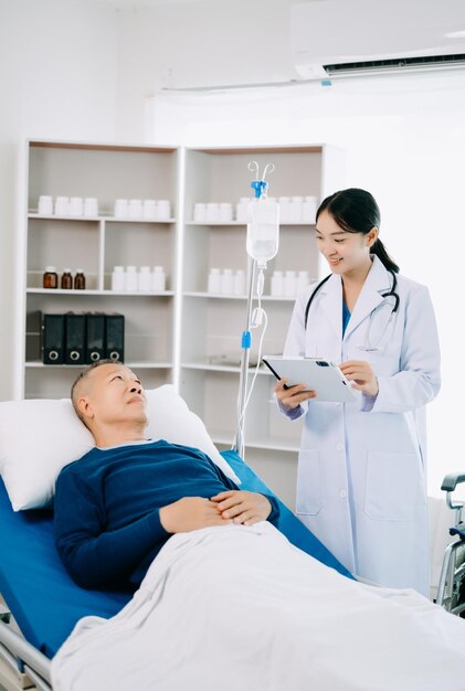 Asian doctor in white suit take notes while discussing and asian elderly man patient who lying on bed with receiving saline solution in hospital or clinic