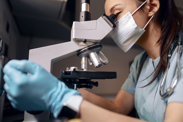 Asian doctor with medical mask and gloves looks into microscope at table in laboratory