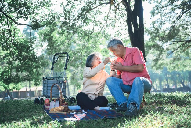 Фото Азиатская пожилая пара привлекательная жена и муж пикник пить чай и отдыхать на зеленой лужайке в саду улыбаются и счастливы вместе к отношениям пенсионного возраста и концепции природы
