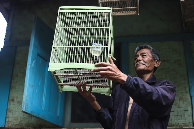 Asian elderly man holding green bird cage outside traditional wooden house