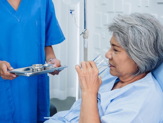 Asian elderly patient taking medicine hold drinking water glass and pill from young nurse in blue scrub suit in hospital room daily medicine or vitamin supplements senior healthcare and medical