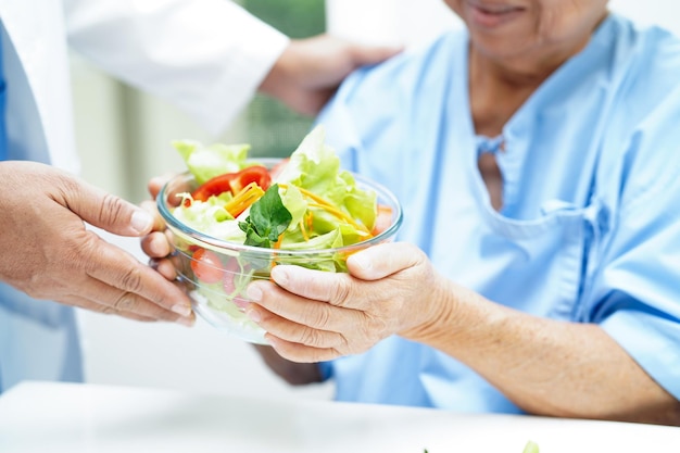 アジアの高齢の女性患者が病院でサルモンステーキと野菜サラダを健康的な食べ物として食べています