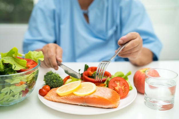 病院で野菜の健康食品とサーモンステーキの朝食を食べるアジアの年配の女性患者