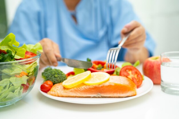 病院で野菜の健康食品とサーモンステーキの朝食を食べるアジアの年配の女性患者