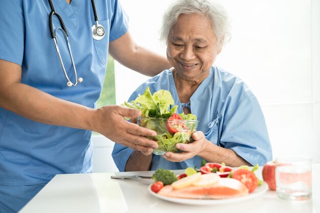 病院で野菜の健康食品とサーモンステーキの朝食を食べるアジアの年配の女性患者