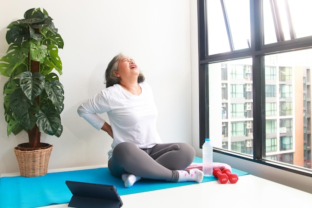 Asian elderly woman sit exercise at home Do yoga poses according to an online fitness teacher via video call via tablet. Social distancing, maintaining the health of the elderly. backache