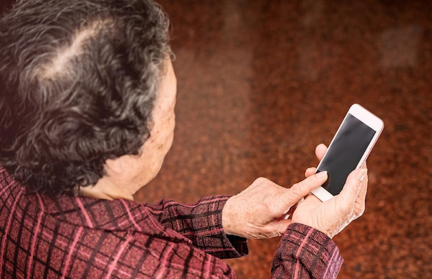 座って現代のスマートフォンで何かを見ているアジアの年配の女性が自宅で他の人とつながる生活技術をクローズアップ