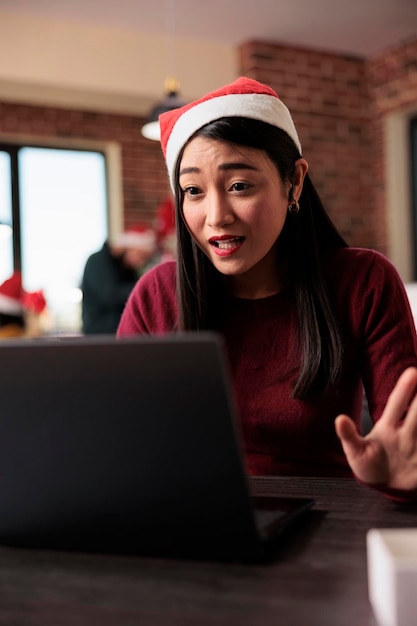 Asian employee attending videocall meeting in office decorated with christmas tree. Talking on online remote videoconference and teleconference call in workplace with holiday ornaments.