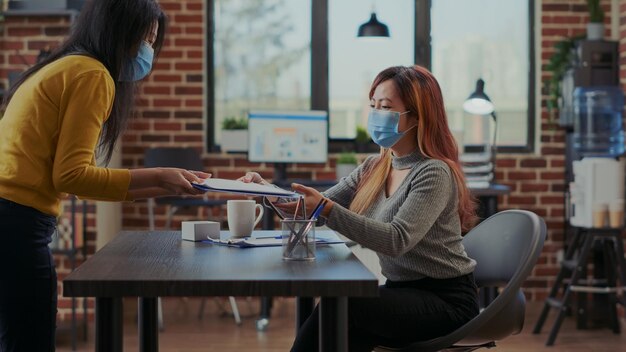 Foto impiegato asiatico con maschera facciale che partecipa a un colloquio di lavoro con il candidato in ufficio. l'operatore delle risorse umane controlla il cv del candidato prima dell'assunzione. reclutamento del lavoro