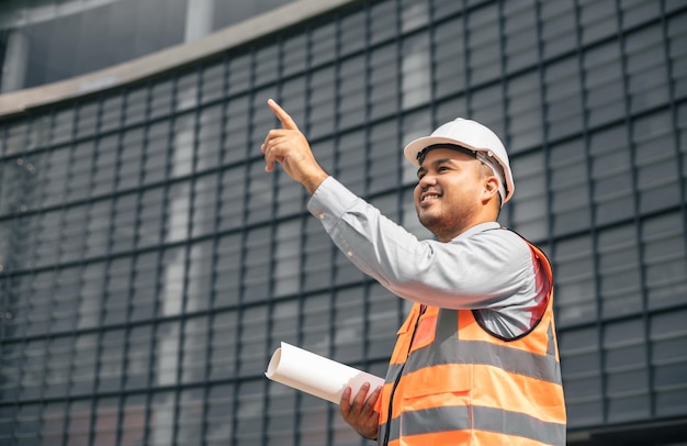 Asian engineer handsome man or architect looking forward holding paperwork blueprint with white safety helmet in construction site Standing at modern building