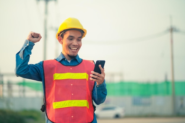 Asian engineer holding smartphone standing outdoor on site\
construction posting confident engineer construction happy job\
success with in target planning