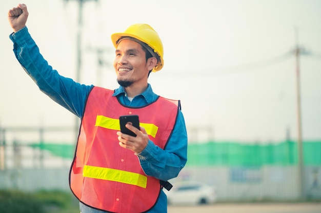 Азиатский инженер держит смартфон, стоящий на открытом воздухе на строительной площадке, уверенно публикует сообщение Инженер-строитель счастливый успех в работе с целевым планированием