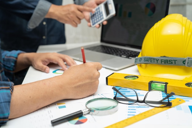 Asian engineer and partner team learn and talk with house plan\
project blueprint graph and yellow helmet and engineering\
construction tools on table work