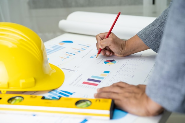 Asian engineer with architectural house plan project blueprint graph and yellow helmet