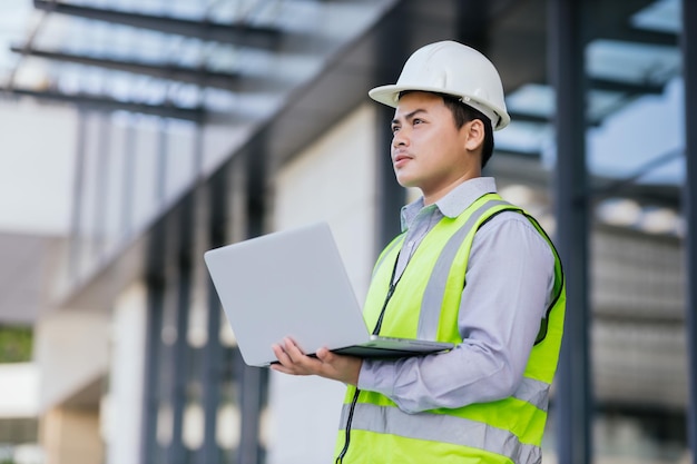 Asian engineer young man wearing safety vest and helmet\
standing using computer for check work on building construction\
site background engineering construction worker concept