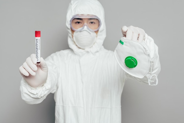 Asian epidemiologist in hazmat suit looking at camera while holding respirator mask and test tube