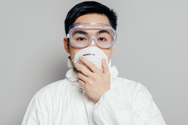 Asian epidemiologist in hazmat suit touching respirator mask while looking at camera isolated on