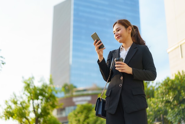 Asian executive donna lavoratrice tenendo la tazza di caffè e utilizzando un telefono cellulare in strada con edifici per uffici in background a bangkok, in thailandia.