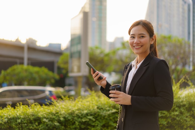 コーヒーカップを持って、タイのバンコクの背景にオフィスビルがある通りで携帯電話を使用しているアジアのエグゼクティブワーキングウーマン。