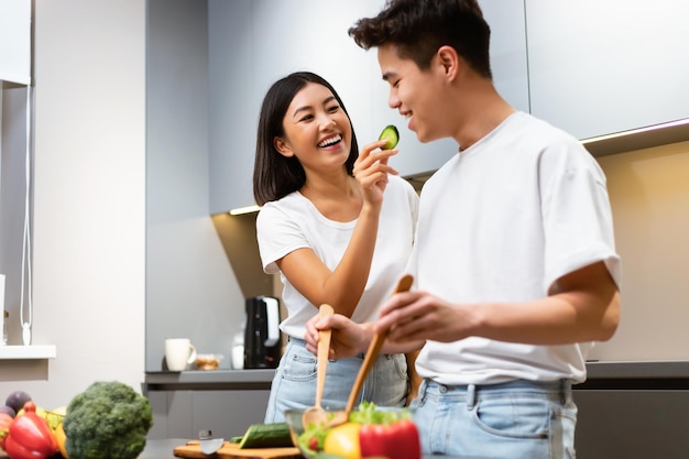 Famiglia asiatica che cucina cibo in cucina. moglie felice che nutre il marito che degusta un pasto che fa l'insalata a casa. coppia coreana divertendosi a preparare la cena insieme nel fine settimana. concetto sano