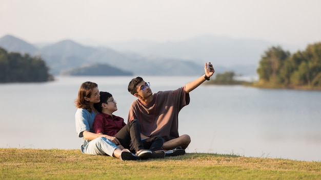 Asian family three members, mother and two young sons, sitting together beside huge lake with mountains and water in background. They using smartphone to take photos.