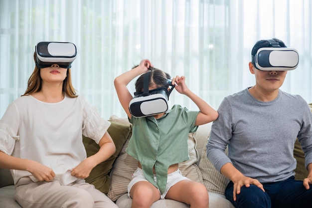 Asian family wear vr glasses headsets sitting on sofa in living room, game entertainment innovation technology, Happy family have mother father and daughter watching movie or playing video game