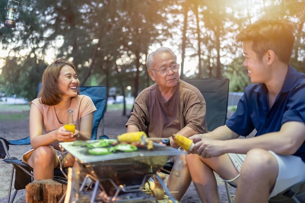 シニアの飲酒と一緒にバーベキューをしているアジアの家族。夏休みのキャンプ中に夕食にグリルバーベキューを調理します。
