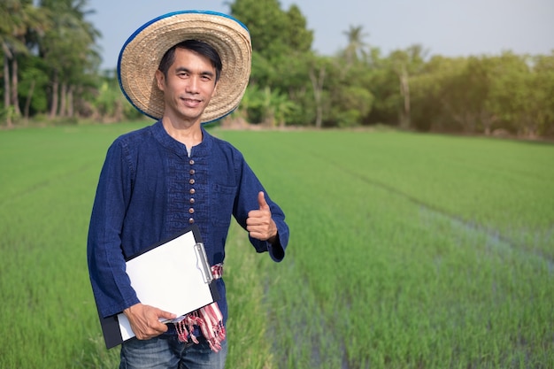 アジアの農家の男性は、伝統的な衣装を着て立って、緑の稲作農場で板紙の笑顔を保持します