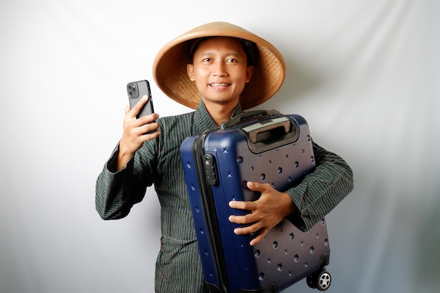 写真 旅行用スーツケースとスマートフォンを背負って幸せに微笑むキャッピングを着たアジアの農夫