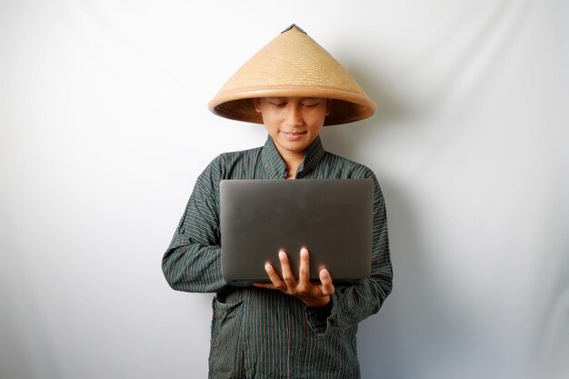 Foto un contadino asiatico che indossa lurik e caping che guarda il portatile con una faccia seria