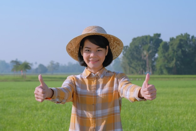 Азиатские женщины-фермеры носят большой палец желтой рубашки и улыбаются, глядя на зеленую ферму.