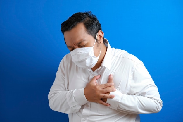 A asian fat guy wearing mask feeling pain in his chest, gesture holding left chest. Blue background