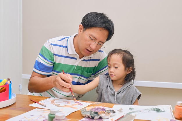 Photo asian father and child having fun painting with paintbrush and watercolors at home