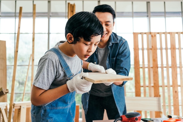 Asian father and son carpentry blowing sawdust from timber plank after sanding to smooth with an electric sander machine together carefully at the home studio carpentry working at workshop studio