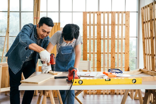 Asian father and son work as a woodworker or carpenter Father teaches his son to drill holes in a wooden plank carefully together with teamwork Craftsman carpentry working at home workshop studio