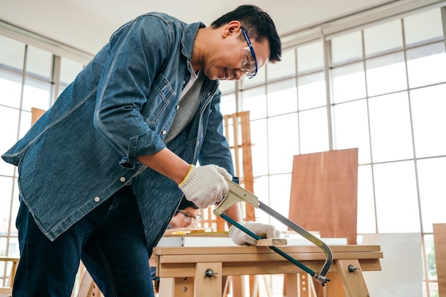 Padre e figlio asiatici lavorano come falegnami o carpentieri il padre indossa occhiali protettivi e ha visto una tavola di legno con una sega da taglio con attenzione falegnameria artigiana che lavora nello studio dell'officina domestica