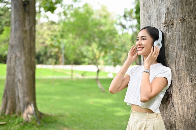 公園の木にもたれ、ヘッドフォンで音楽を聴くアジアの女性