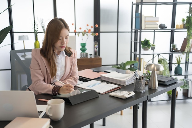 An Asian female businesswoman is seated at her desk writing notes on a tablet while surrounded by papers and a laptop The office setting is contemporary and wellorganized