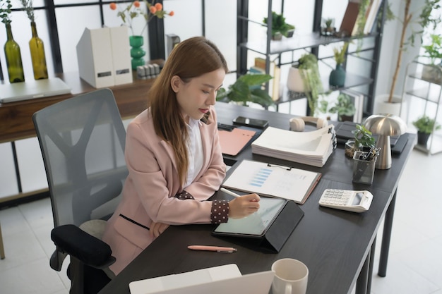 An Asian female businesswoman is seated at her desk writing notes on a tablet while surrounded by papers and a laptop The office setting is contemporary and wellorganized