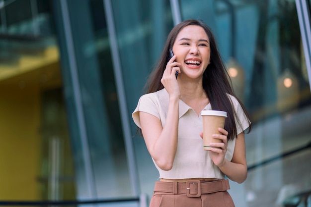 Азиатская женщина вскользь расслабляется, цифровой кочевник-программист смотрит разговор на смартфоне, гуляя по городу с чашкой кофе, держа улыбаясь, веселое позитивное чувство, понижая образ жизни