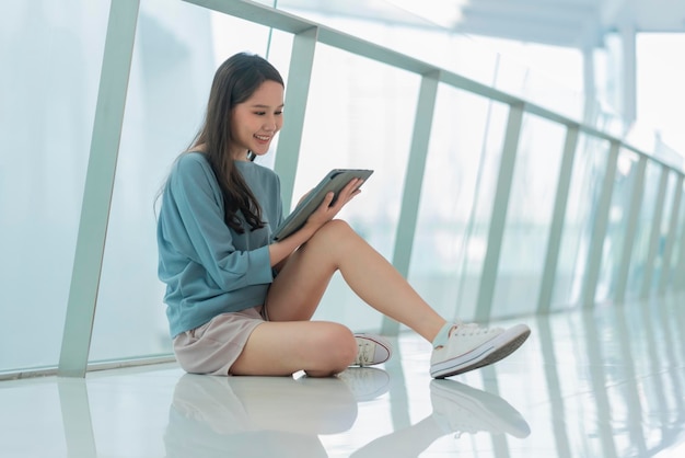 Asian female collage young campus woman sitting on the mall\
floor using tablet browing surf data online social media with\
cheerful fun smilingasian woman digital nomad sit on floor casual\
work online