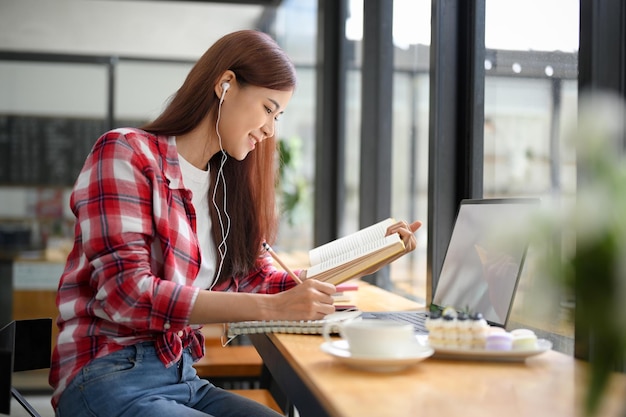 Studentessa di college asiatica che fa i compiti leggendo un libro di testo e ascoltando la lezione online