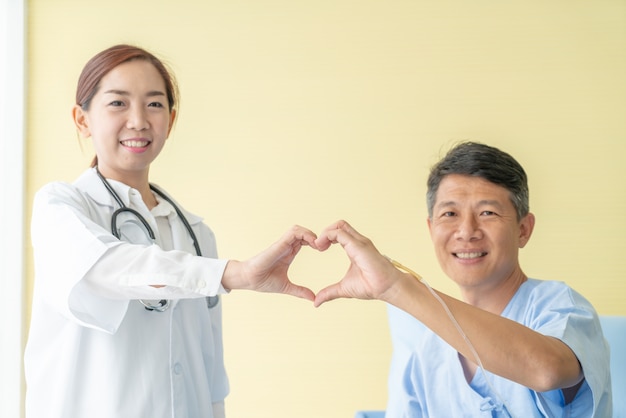 Il medico femminile asiatico che sorride e fa il cuore si rade a mano con il paziente senior