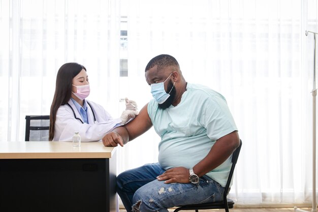 An Asian female doctor wearing a mask and gloves is vaccinating an African-American man against the coronavirus. To build immunity. Hospital medical service concept