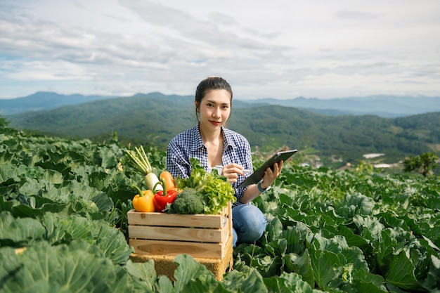 Asian female farmer working early on farm holding wood basket of fresh vegetables and tabletxA