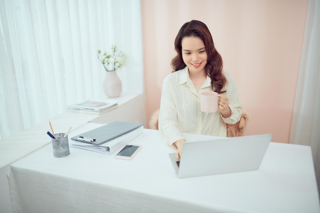 Libero professionista femminile asiatico che si rilassa seduto con una tazza di caffè davanti al computer portatile