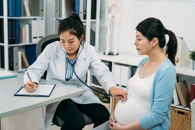 사진 아시아 여성 산부인과 의사는 병원에서 임신을 확인하는 동안 청진기로 임산부를 검사하고 클립보드에 글을 쓰고 있습니다.