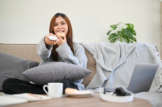 Una donna asiatica nel suo soggiorno si diverte a mangiare le ciambelle mentre guarda uno spettacolo televisivo