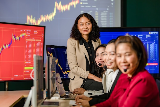 アジアの女性プロ証券取引所マネージャーが机の上に座ってフロントモニターで笑顔カメラを見て